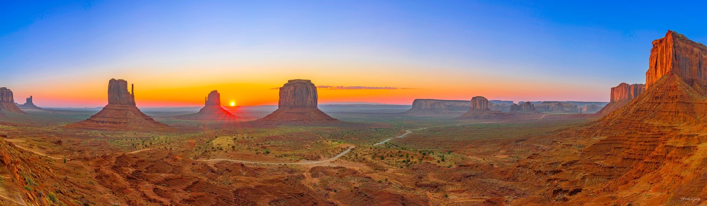 Red rock buttes rise above a desert valley with winding roads, beneath a low sun and an orange-to-blue sky.
