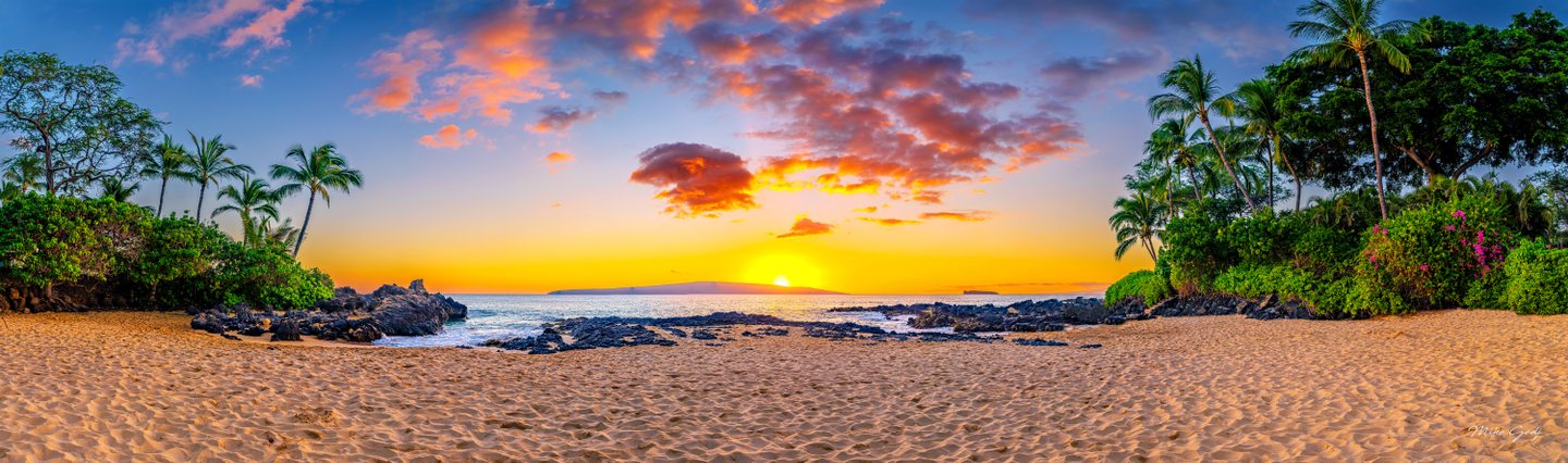 Palm trees frame a sandy beach and dark coastal rocks beneath orange clouds and a low sun over distant land.