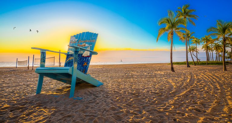 An oversized blue chair marked Fort Lauderdale Beach stands on sand beside palms, beach nets, and a golden sea.