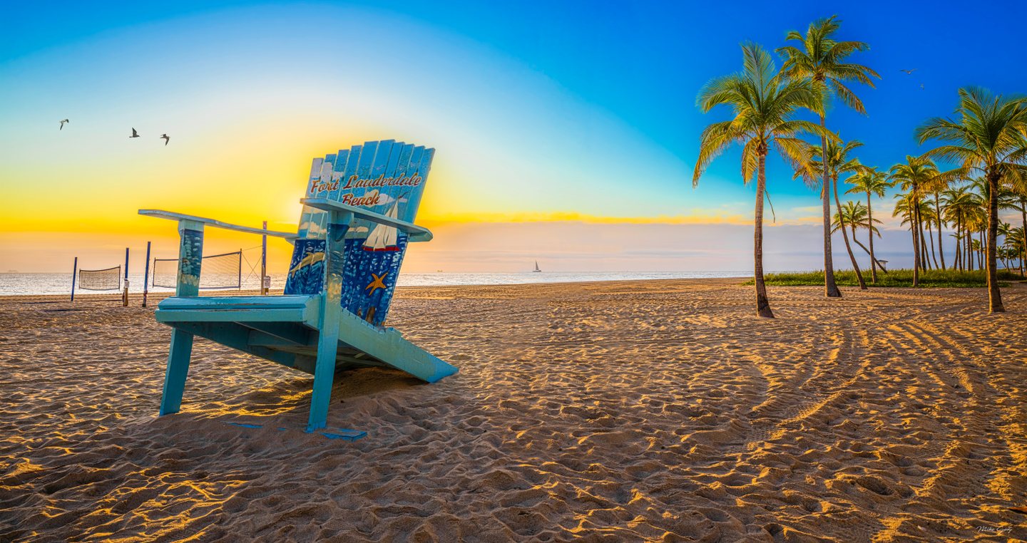 An oversized blue chair marked Fort Lauderdale Beach stands on sand beside palms, beach nets, and a golden sea.