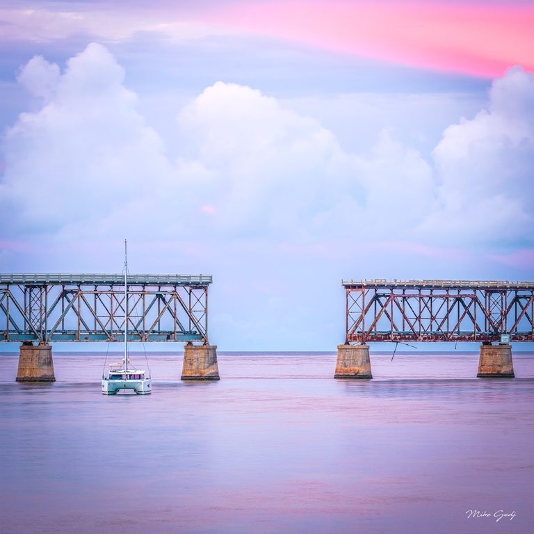 A white catamaran floats before rusted bridge spans with a wide gap, beneath pink and blue clouds.