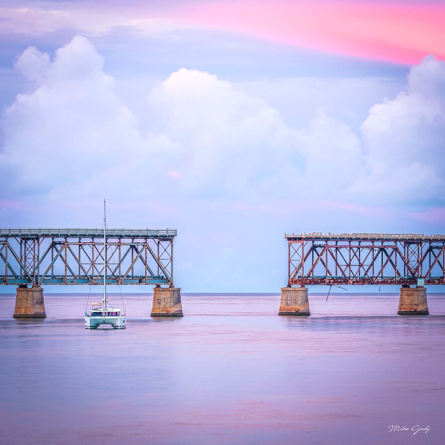 A white catamaran floats before rusted bridge spans with a wide gap, beneath pink and blue clouds.