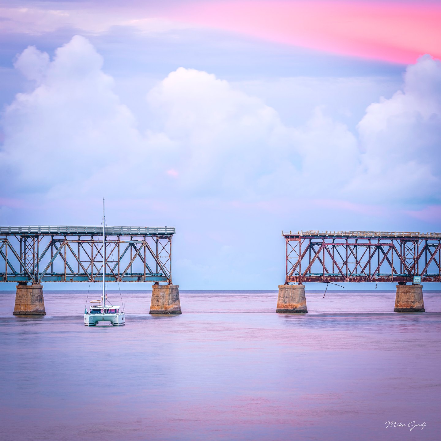 A white catamaran floats before rusted bridge spans with a wide gap, beneath pink and blue clouds.