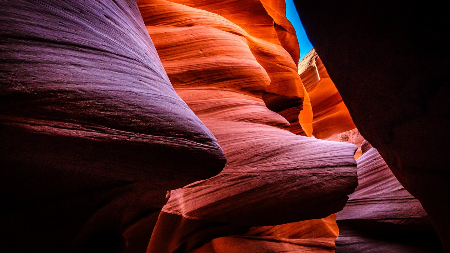 Curving sandstone canyon walls glow orange and violet around deep shadows and a narrow sliver of blue sky.