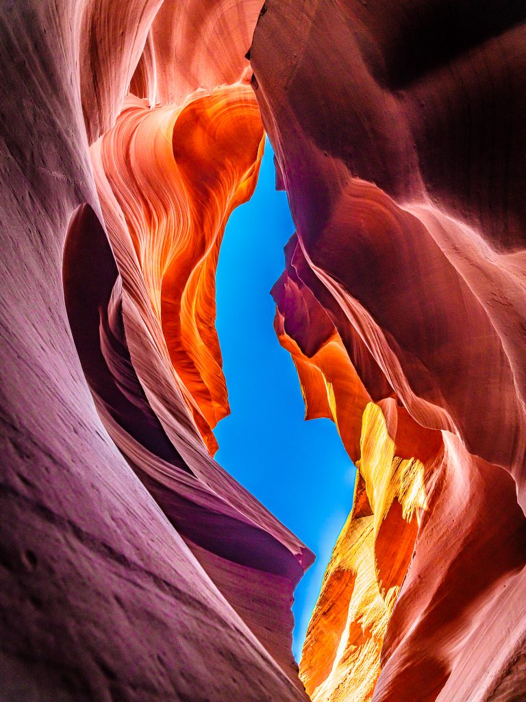 An upward view between rippled red canyon walls reveals a narrow ribbon of vivid blue sky.