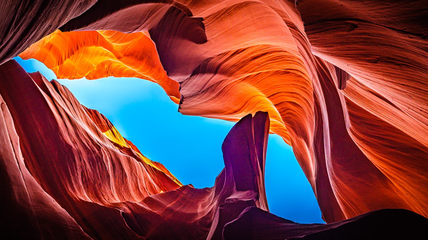 An upward view between rippled red-orange canyon walls framing a winding opening of vivid blue sky.