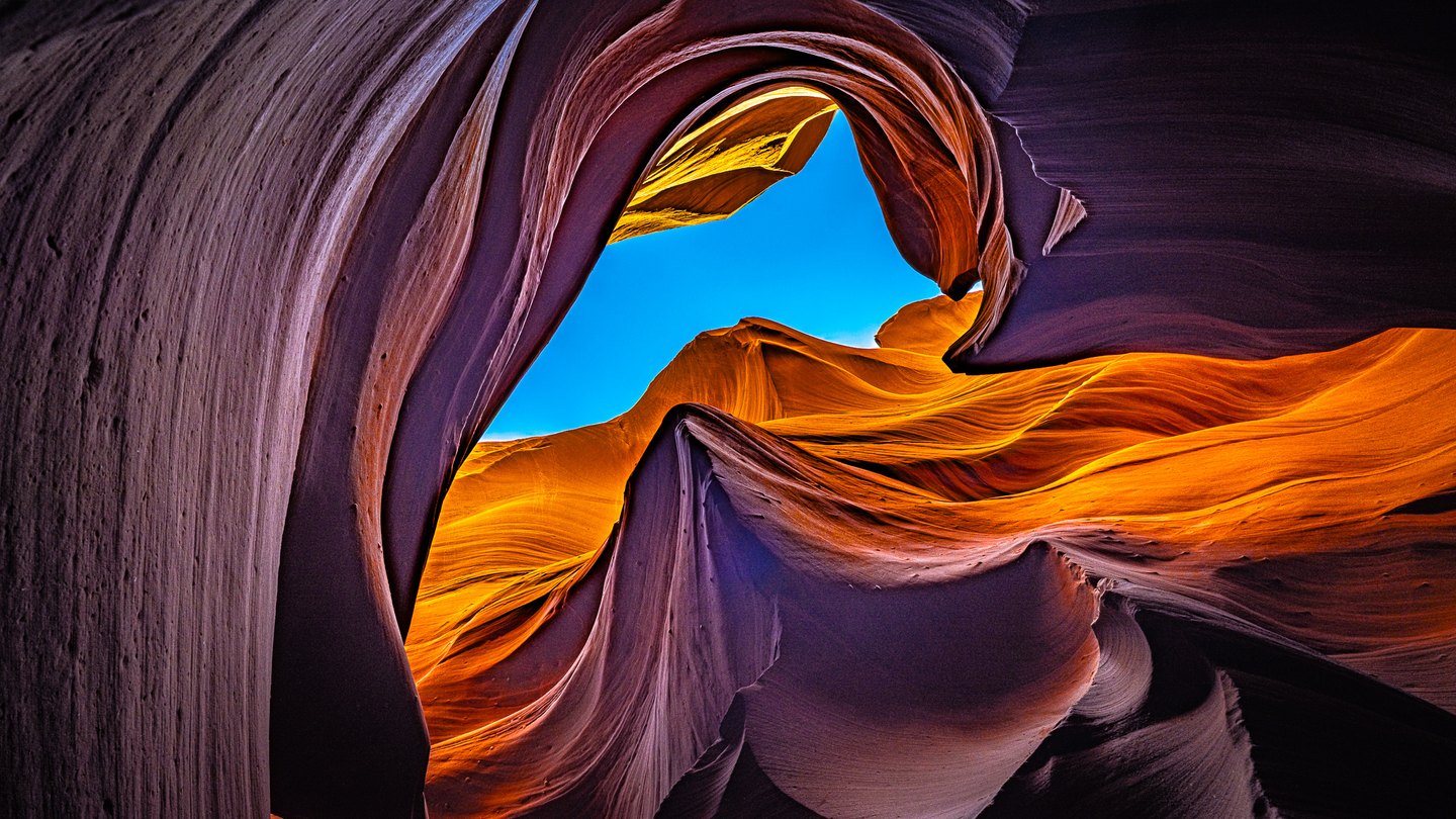 Curving sandstone canyon walls glow orange above purple shadows, framing a bright blue opening of sky.