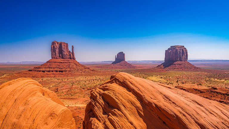 Three red rock buttes rise beyond curved, striated orange sandstone and a sparse desert plain beneath a blue sky.