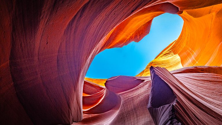 Curving red and purple sandstone walls frame bright blue sky, with sunlight turning the upper edges golden orange.