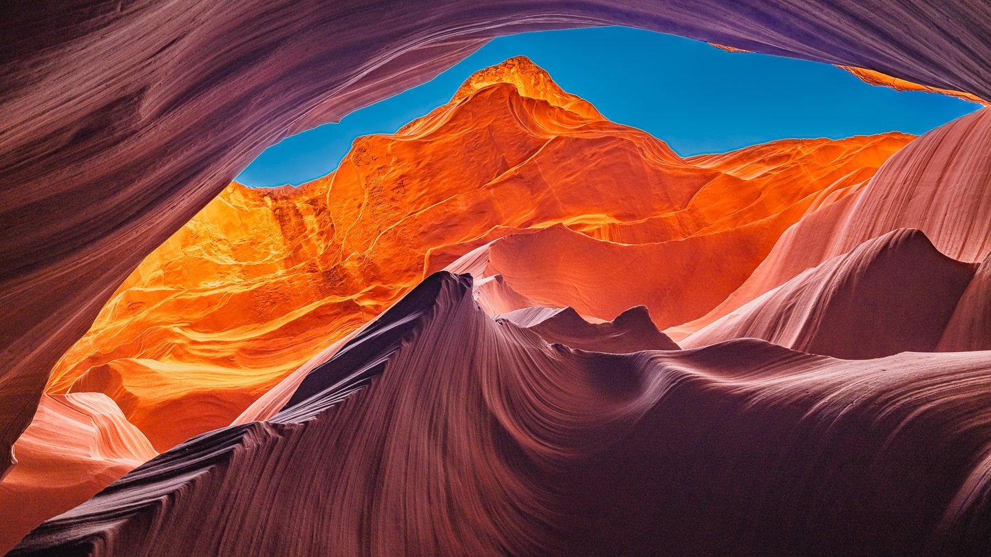 Rippling canyon walls in orange and purple frame a narrow opening of bright blue sky.