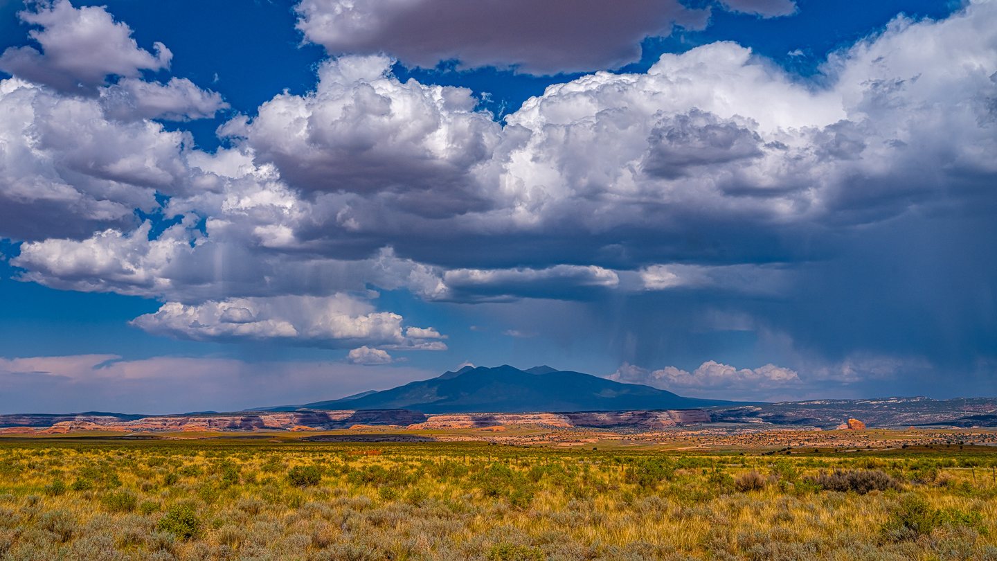 Golden scrubland and orange cliffs stretch toward a dark mountain beneath towering clouds and distant rain.