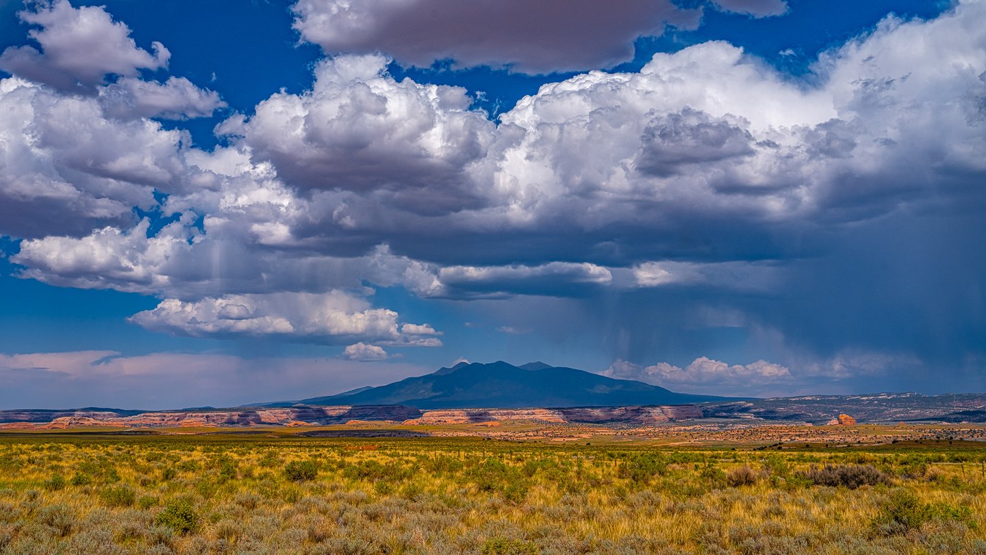 Golden scrubland and orange cliffs stretch toward a dark mountain beneath towering clouds and distant rain.