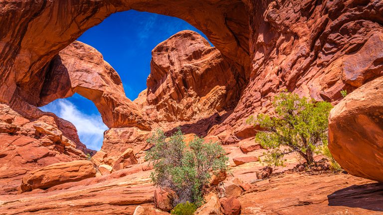 Red rock arches frame blue sky and wispy clouds above sloping stone, scattered boulders, and green shrubs.