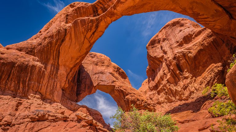 Two red rock arches frame blue sky and wispy clouds above rugged slopes dotted with green shrubs.