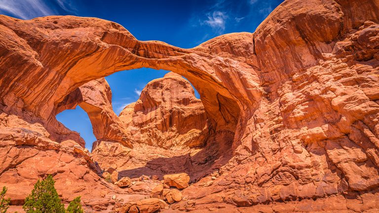 Immense orange rock arches frame blue sky above tiny hikers, scattered boulders, and sparse green shrubs.
