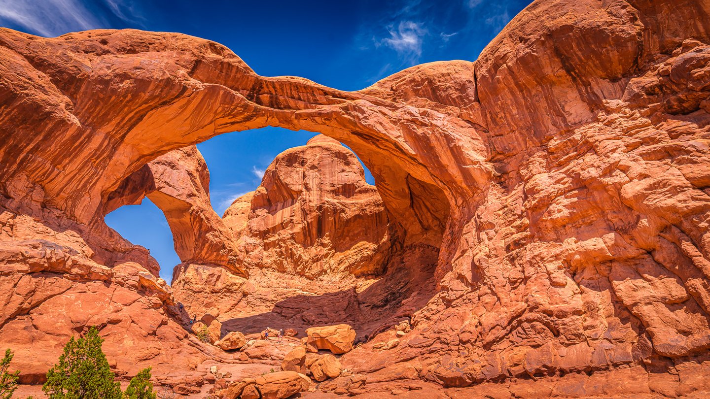 Immense orange rock arches frame blue sky above tiny hikers, scattered boulders, and sparse green shrubs.