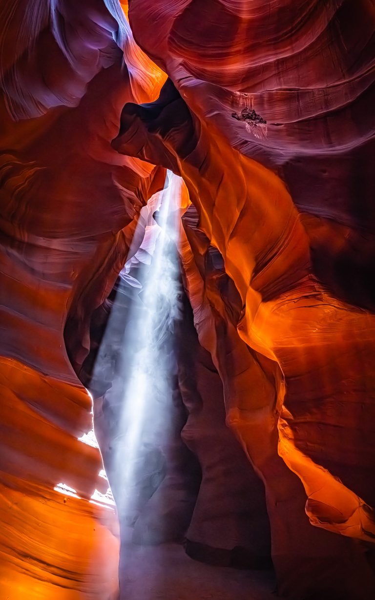 A white shaft of sunlight falls between curving slot canyon walls glowing orange, red, and violet.