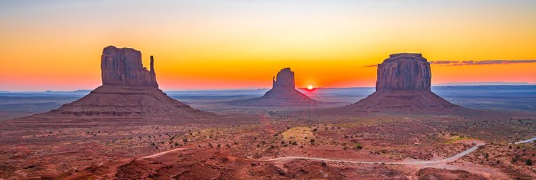 Three towering desert buttes rise above red terrain beneath an orange sky, with the sun low on the horizon.