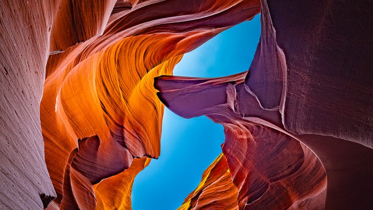 An upward view between curving sandstone walls, glowing orange and shaded purple, frames a narrow blue sky.