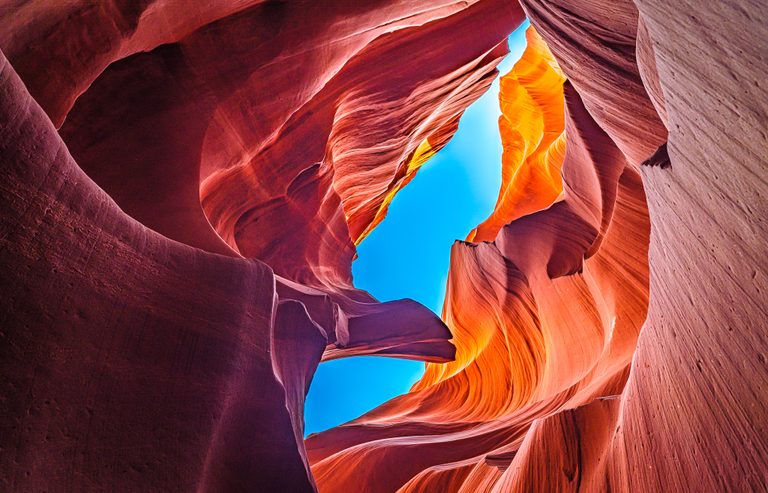 An upward view of curved red sandstone canyon walls framing narrow openings of vivid blue sky.