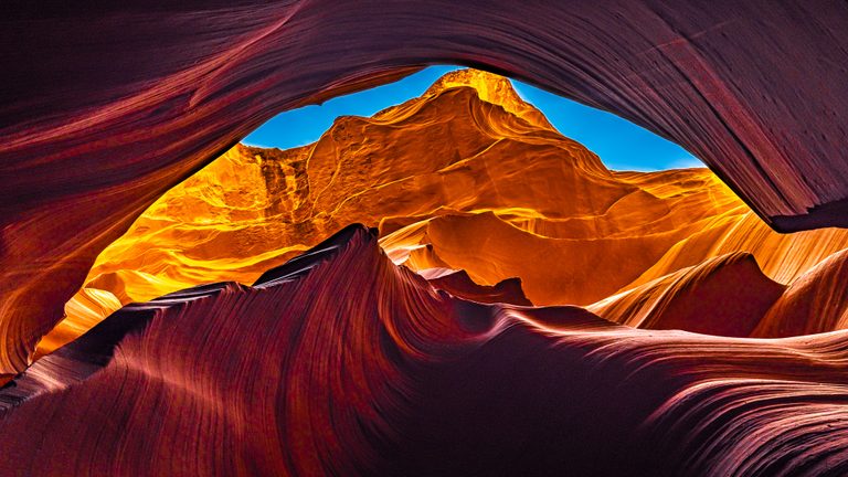 Curving red canyon walls frame sunlit orange rock and a narrow opening of vivid blue sky.