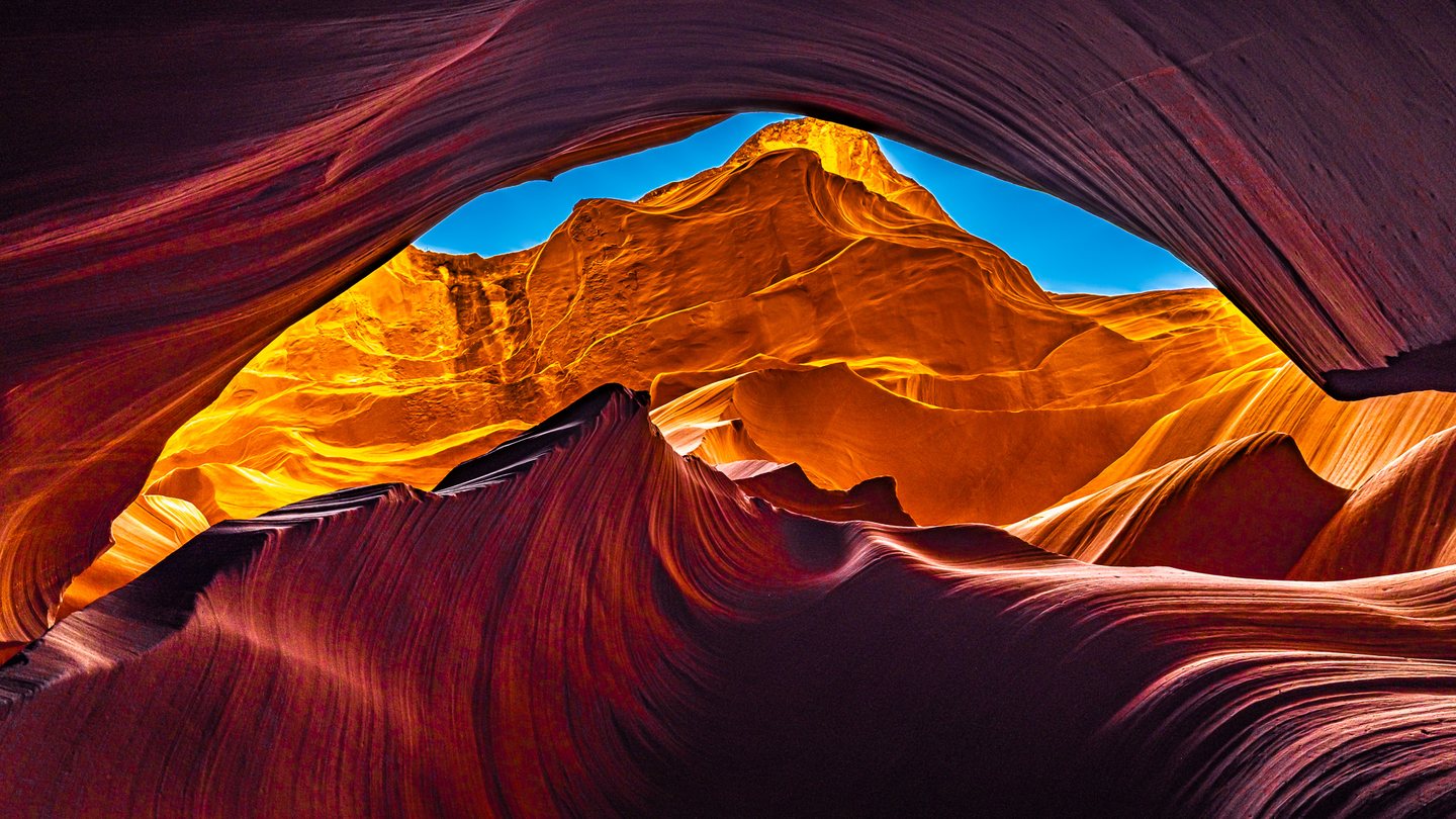 Curving red canyon walls frame sunlit orange rock and a narrow opening of vivid blue sky.