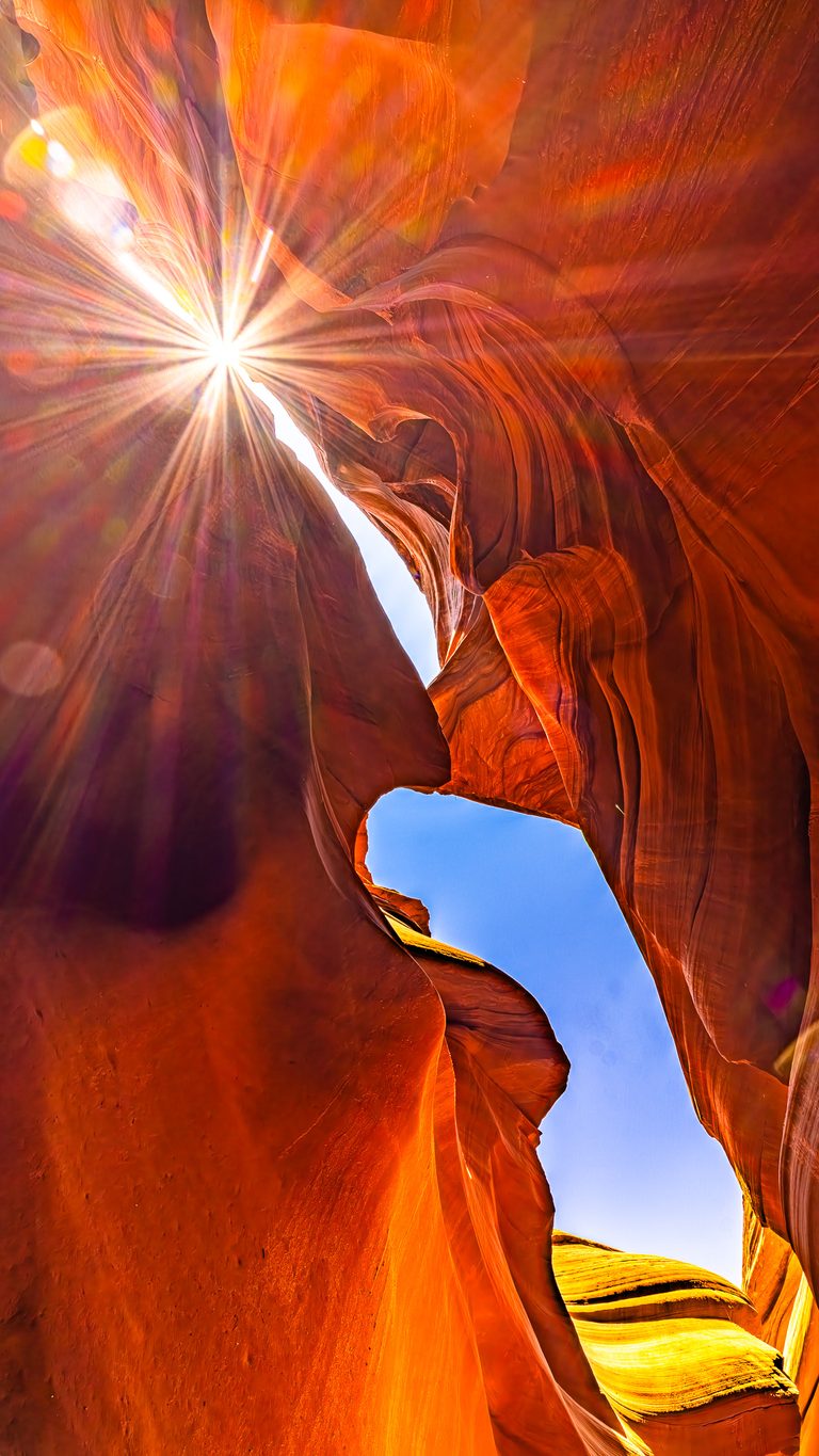A sunburst shines between curved orange sandstone walls, framing narrow openings of blue sky in a slot canyon.