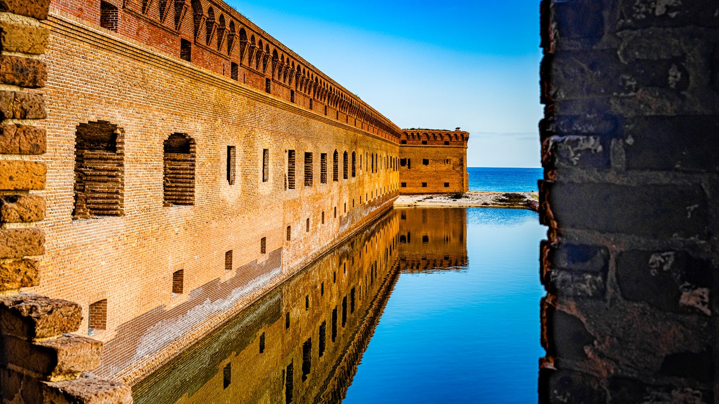 Sunlit brick fort walls reflect in still water, framed by foreground masonry, with blue sea and sky beyond.