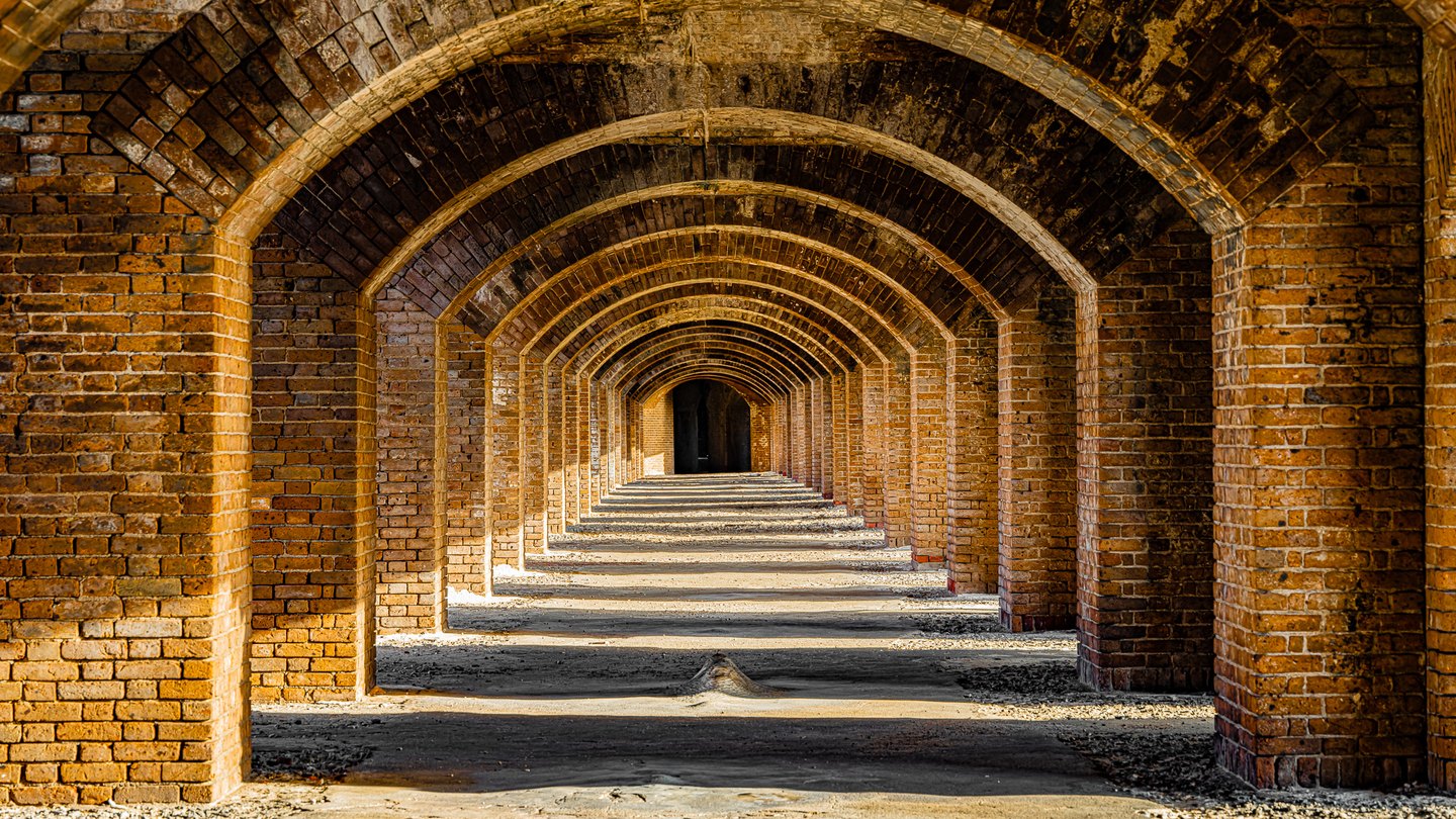Repeated brick arches recede toward a dark opening, with bands of sunlight and shadow crossing the rough floor.