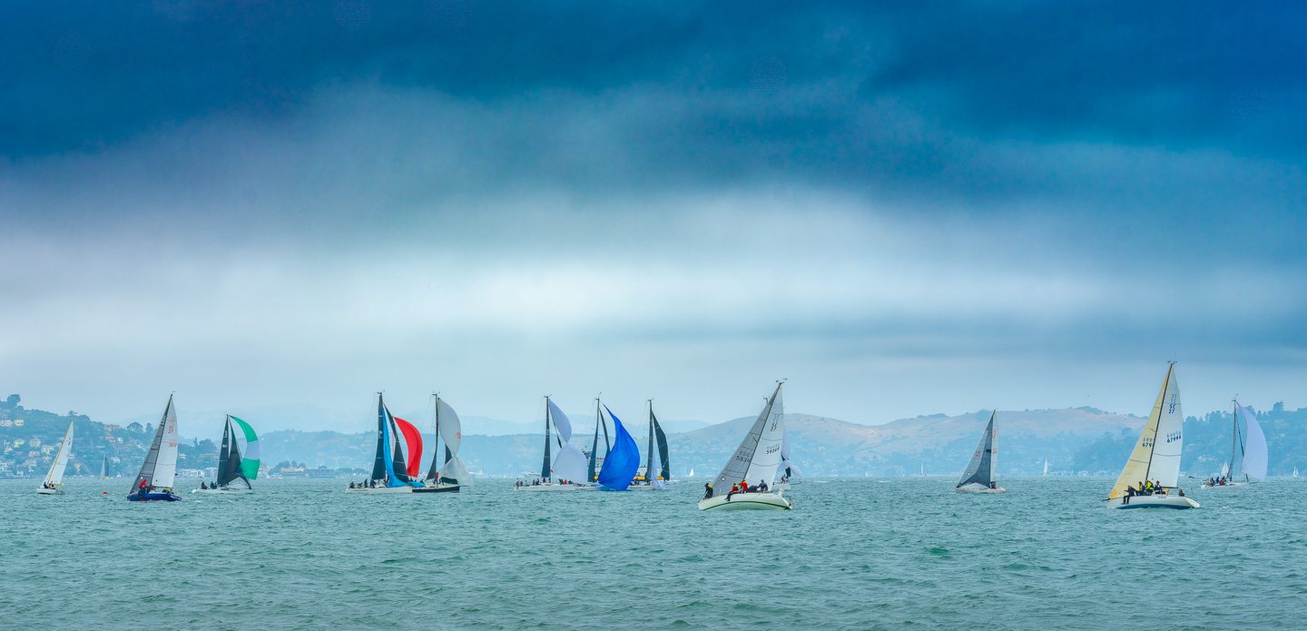 Sailboats with colorful sails cross rippled blue-green water beneath a cloudy sky, with hills in the distance.