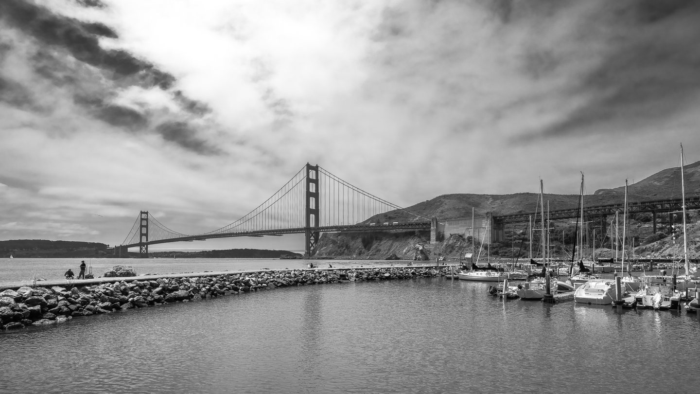 Black-and-white view of a suspension bridge beyond a rocky breakwater, with moored sailboats beneath a cloudy sky.