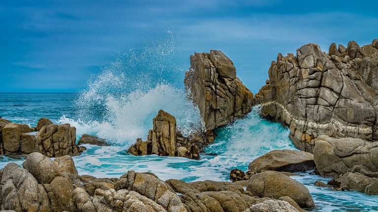 White surf bursts above jagged coastal rocks as turquoise water swirls beneath a blue, lightly clouded sky.