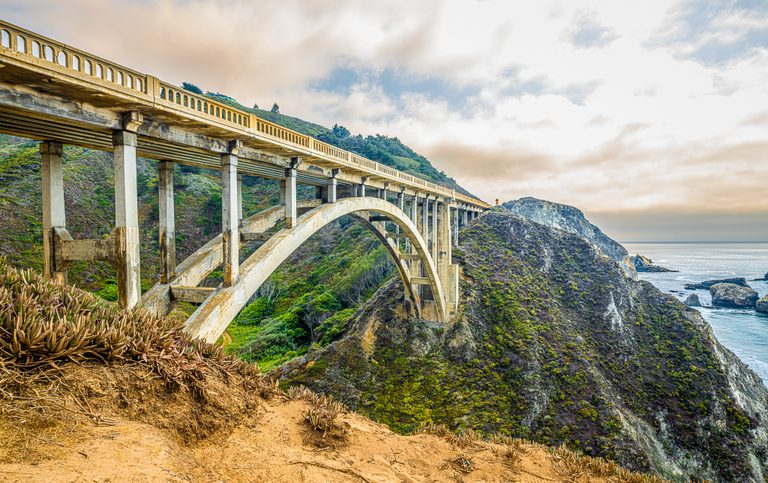 A concrete arch bridge spans a green coastal ravine beside rocky cliffs and ocean beneath a cloudy sky.