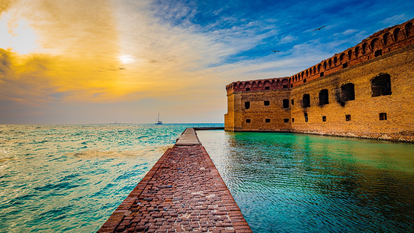 A brick walkway runs beside a coastal fort above turquoise water, with a sailboat and birds beneath a golden-blue sky.