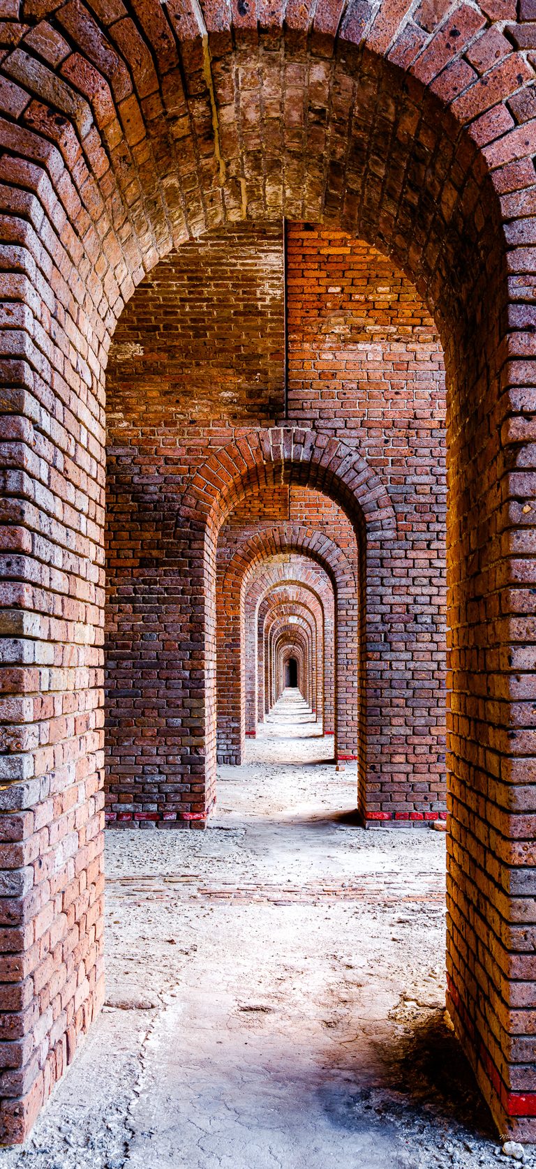 A long sequence of red brick arches recedes toward a dark doorway above a pale, uneven floor.