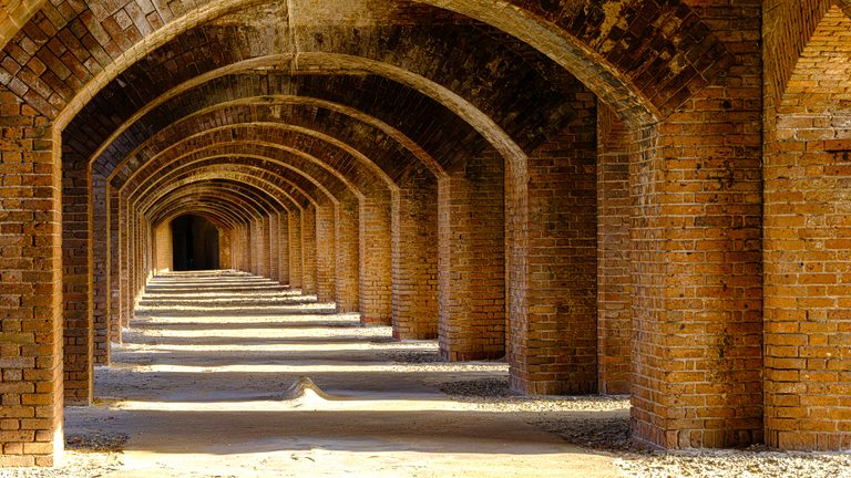 Repeating brick arches recede toward a dark opening, with bands of sunlight crossing the rough floor.