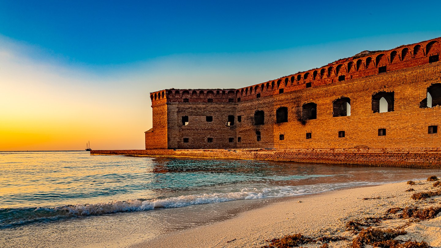 Golden light illuminates a brick coastal fort beside gentle waves, a sandy beach, and a distant sailboat.