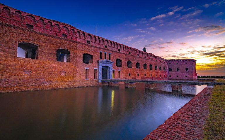 A red brick fort and entrance bridge stand above a reflective moat beneath a blue and gold twilight sky.