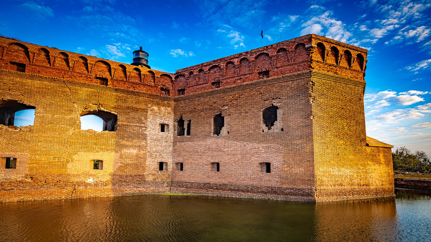 Sunlit brick fort walls rise above rippling water, with a lantern tower and a bird against a cloud-streaked blue sky.