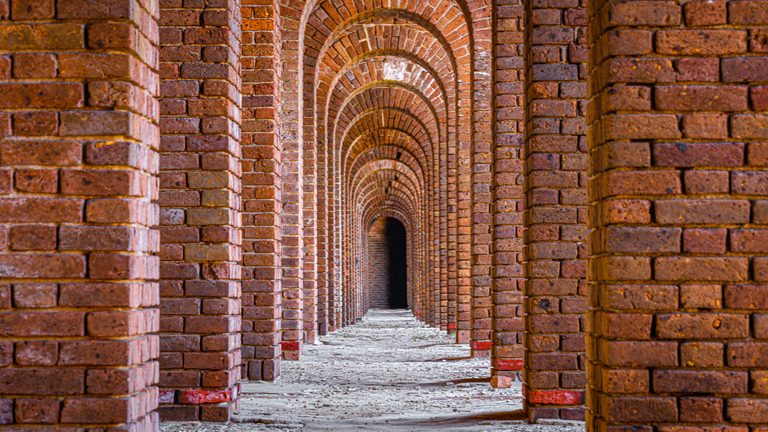 Rows of reddish brick arches recede over a pale, uneven floor toward a dark opening.