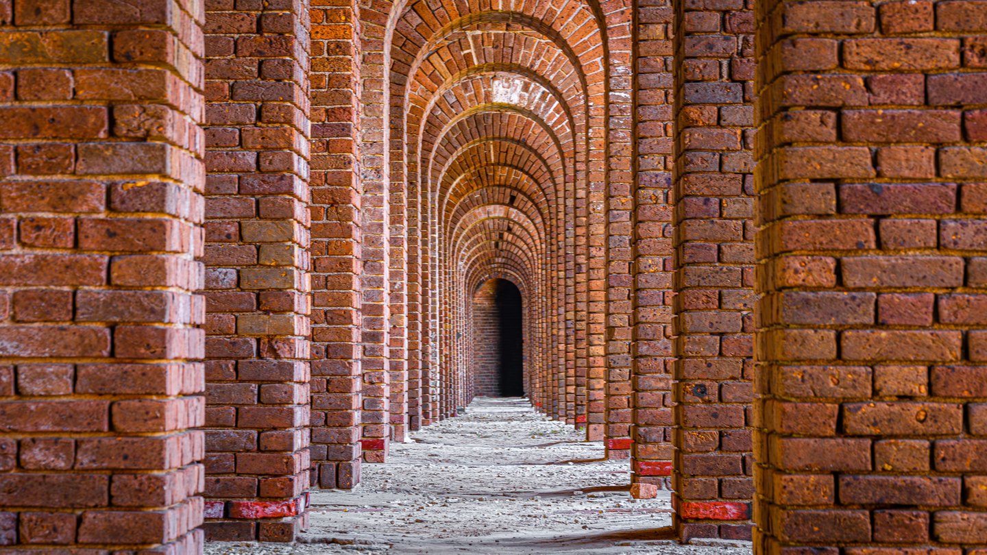 Rows of reddish brick arches recede over a pale, uneven floor toward a dark opening.
