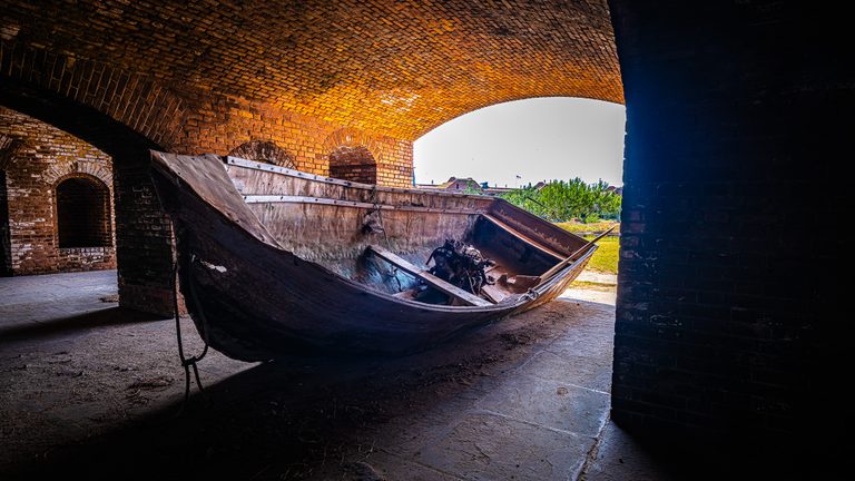 A weathered boat with an exposed engine rests beneath sunlit brick vaults beside a bright courtyard.