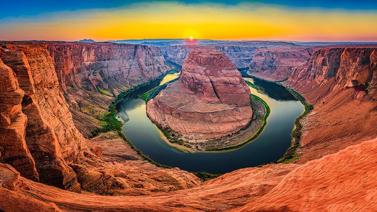 A river curves around a massive red rock formation beneath steep canyon walls and a golden sky with a low sun.