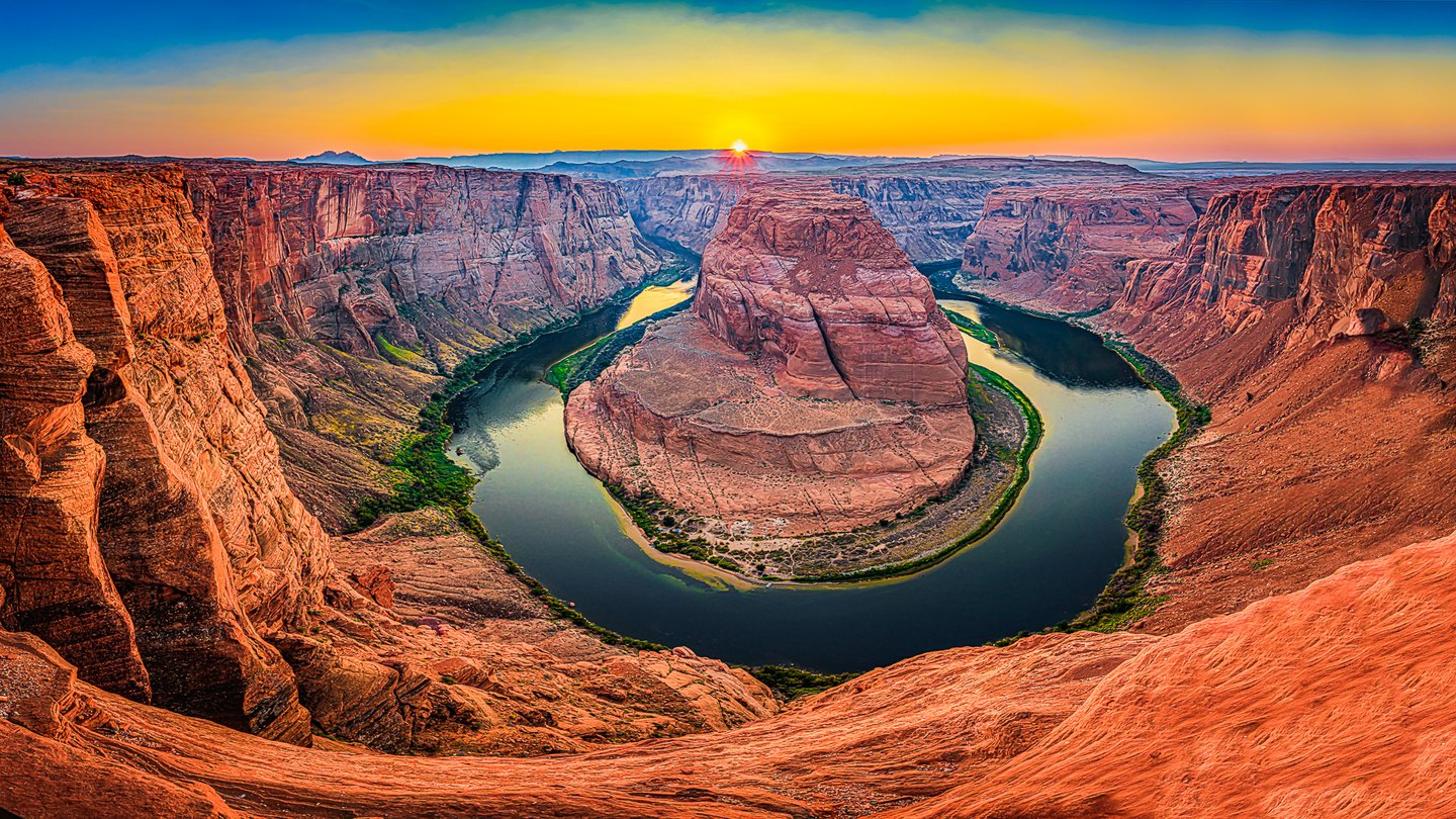A river curves around a massive red rock formation beneath steep canyon walls and a golden sky with a low sun.