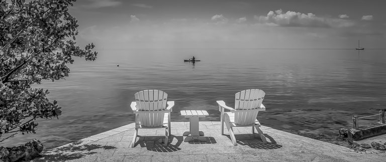 Two white chairs and a small table face calm water, with a paddler offshore and a distant sailboat in black and white.