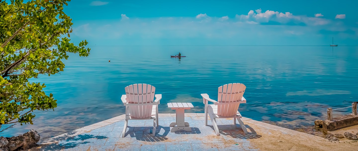Two pale Adirondack chairs and a small table face calm turquoise water, with a paddler and distant sailboat.
