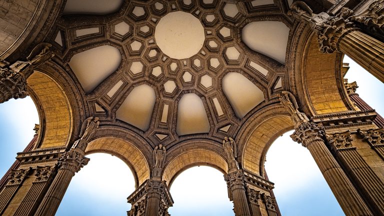 Upward view of an ornate coffered dome, sculpted figures, and fluted columns framing arches of pale blue sky.