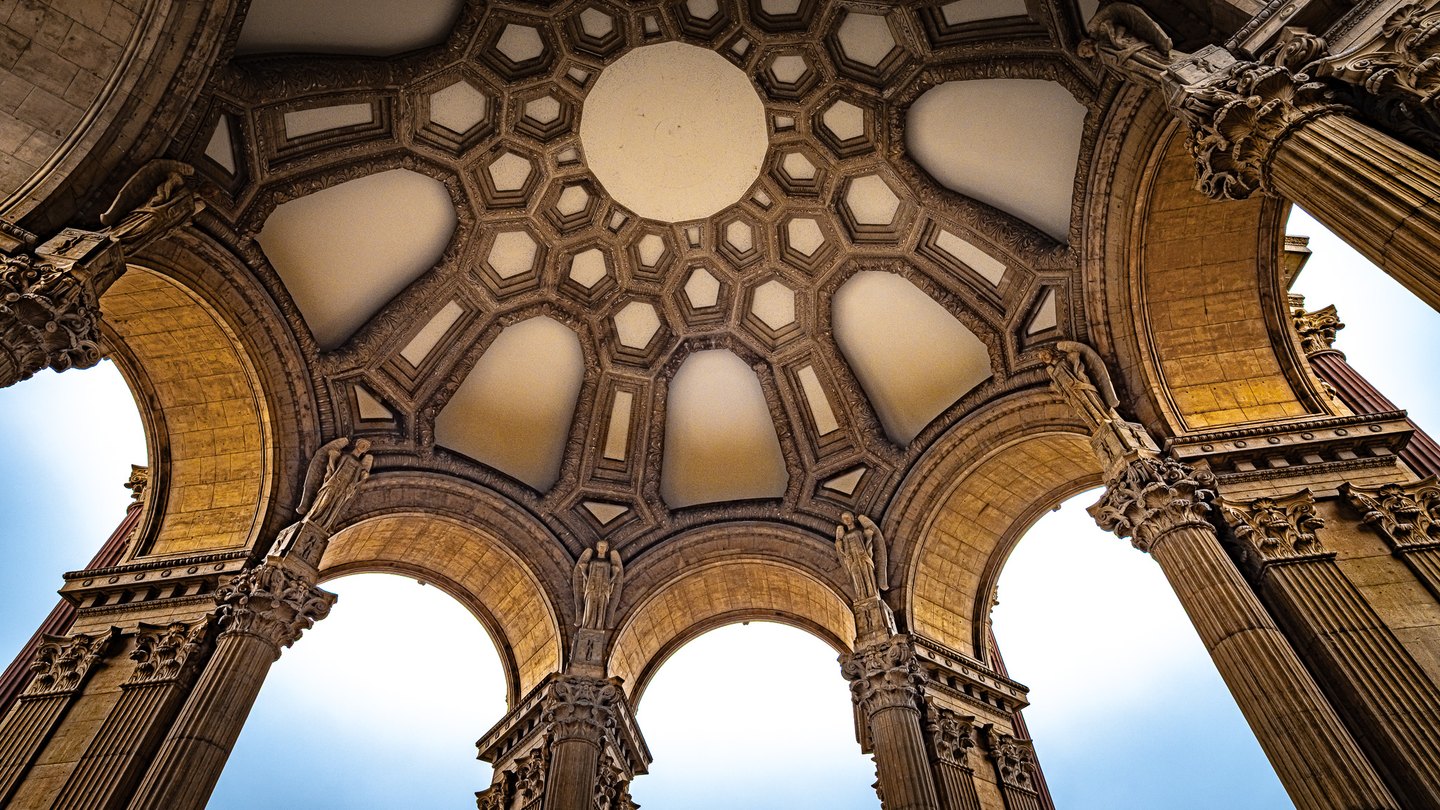 Upward view of an ornate coffered dome, sculpted figures, and fluted columns framing arches of pale blue sky.
