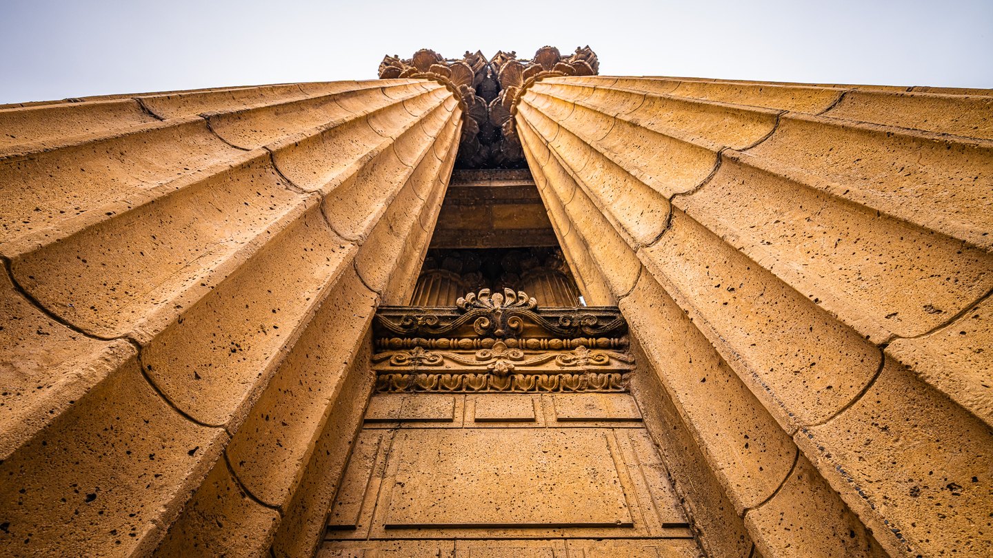 Low-angle view of two golden fluted stone columns with ornate capitals and carved details against a pale sky.