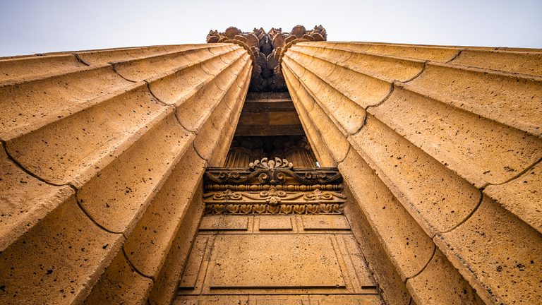 Low-angle view of two golden fluted stone columns with ornate capitals and carved details against a pale sky.